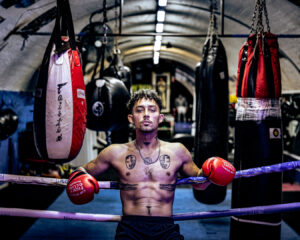 Shirtless male boxer wearing red gloves resting between rounds in a gritty Melbourne boxing gym.