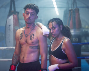Female boxer with wrapped hands leaning on male boxer wearing red gloves, both posing confidently in boxing ring at gritty Melbourne gym.