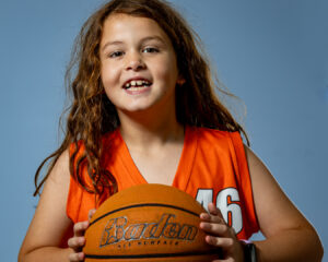 Portrait of a smiling young girl in a basketball jersey holding a ball, captured in a youth sports photography session.