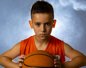 Focused young basketball player in an orange jersey holding a ball, lit dramatically with a smoky studio backdrop.