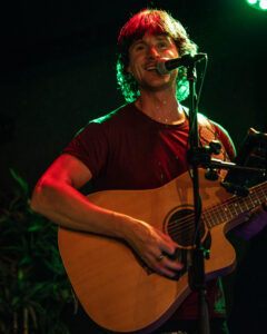 Lead singer playing acoustic guitar, standing at microphone, smiling and looking at camera
