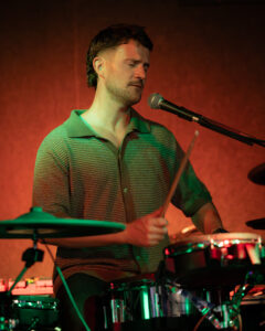 Drummer with eyes closed singing into microphone behind drum kit, holding drumstick