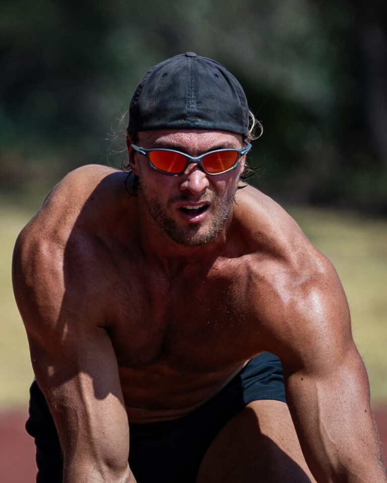 A shirtless athlete wearing reflective sunglasses crouches on the track during an intense sprint training session.