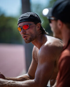 An athlete wearing reflective sunglasses rests and talks during a training session on an outdoor athletics track.