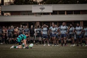A rugby player kneels to prepare a conversion kick while opposition players watch from the background after a match-winning moment.