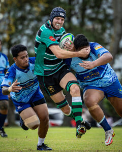 A rugby player in a green and black jersey fends off a tackle while charging through two defenders during a match.