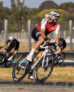 A cyclist in a red and white kit leans through a corner during a criterium race while riders trail behind in the background.