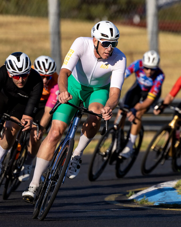 A cyclist in an Australian kit leans aggressively into a corner during a criterium race while the peloton follows closely behind.
