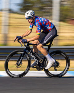 A cyclist in a blue and red jersey races along the circuit at speed, captured with a panning motion blur background.