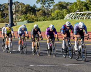 A group of cyclists ride in close formation down the main straight during a criterium race under bright afternoon light.